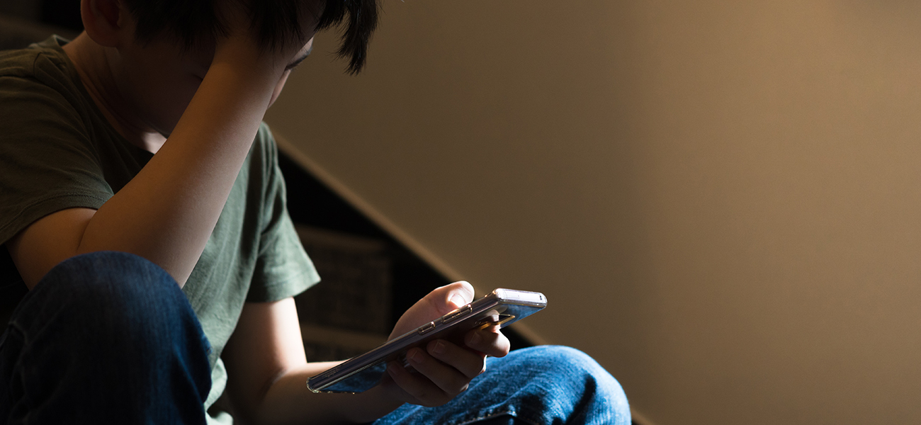 Teenage boy sits on stairs covering his face with his hand while other hand holds a phone